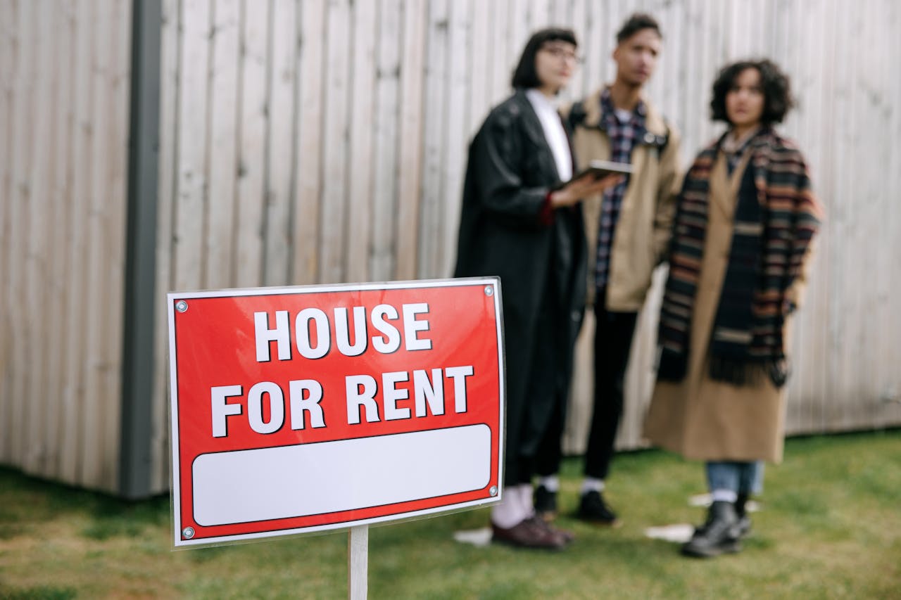 Red and white sign reading ‘HOUSE FOR RENT’ in front of a wooden fence with three people standing blurred in the background.