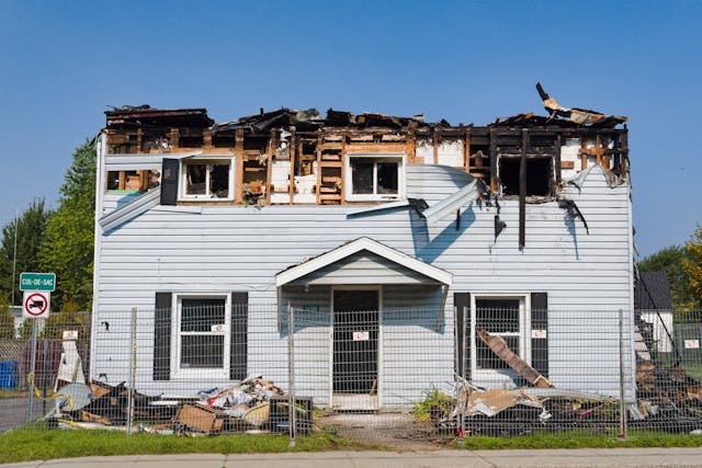 Two-story house severely damaged by fire with charred upper floor and debris around it, fenced off on a clear day.