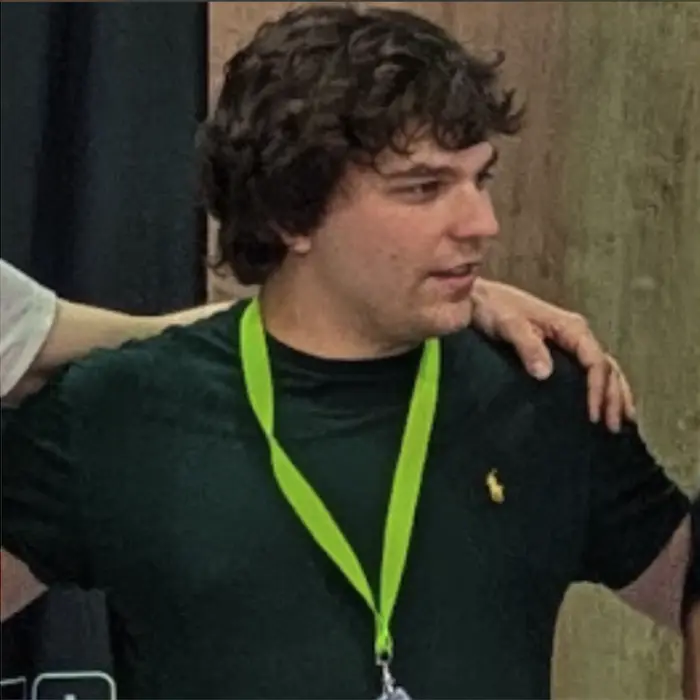 Young man with curly dark hair wearing a black shirt and a green lanyard, with a hand resting on his shoulder.