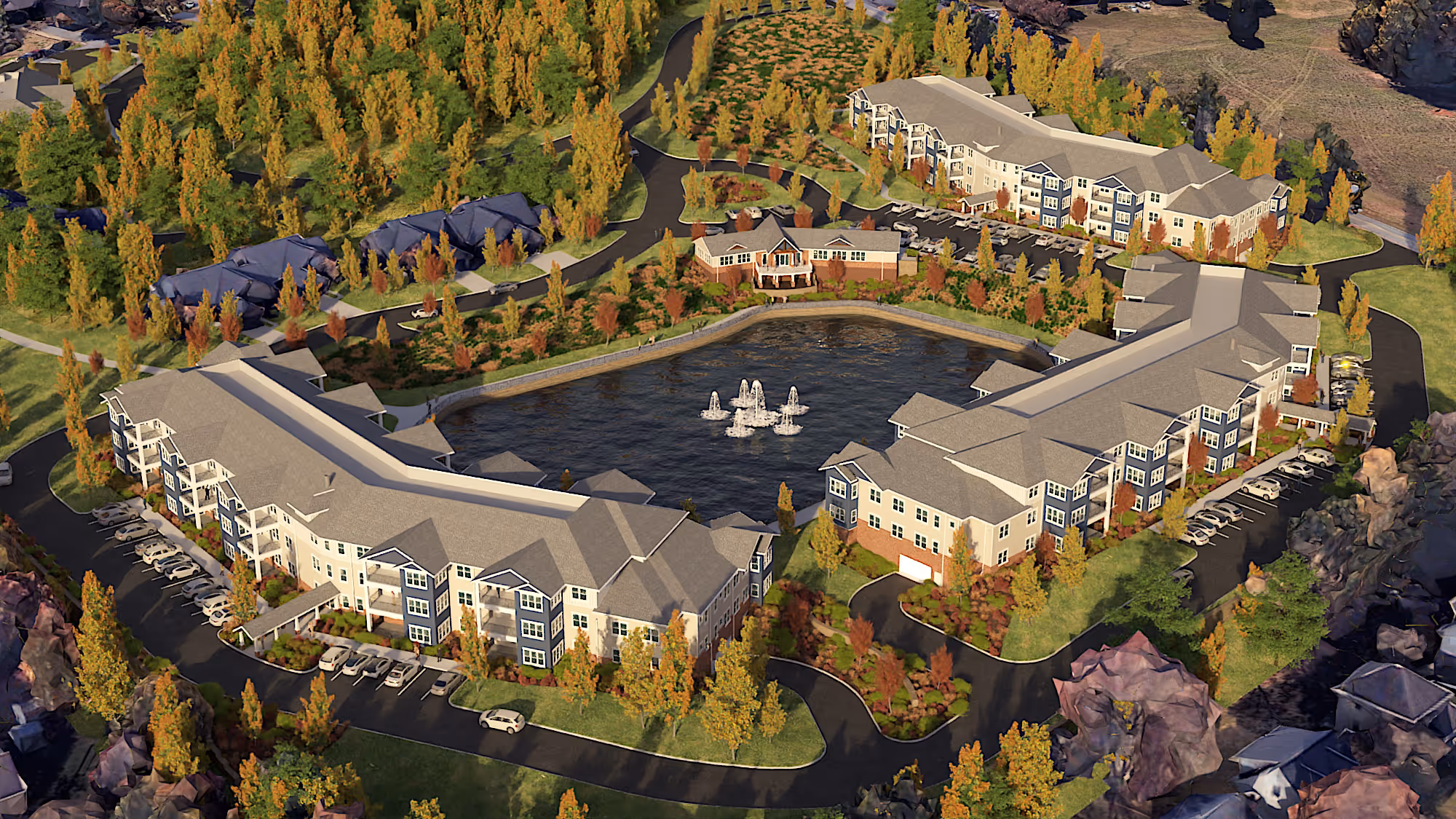 Aerial view of a residential complex with two large apartment buildings surrounding a pond with fountains, surrounded by trees with autumn foliage.