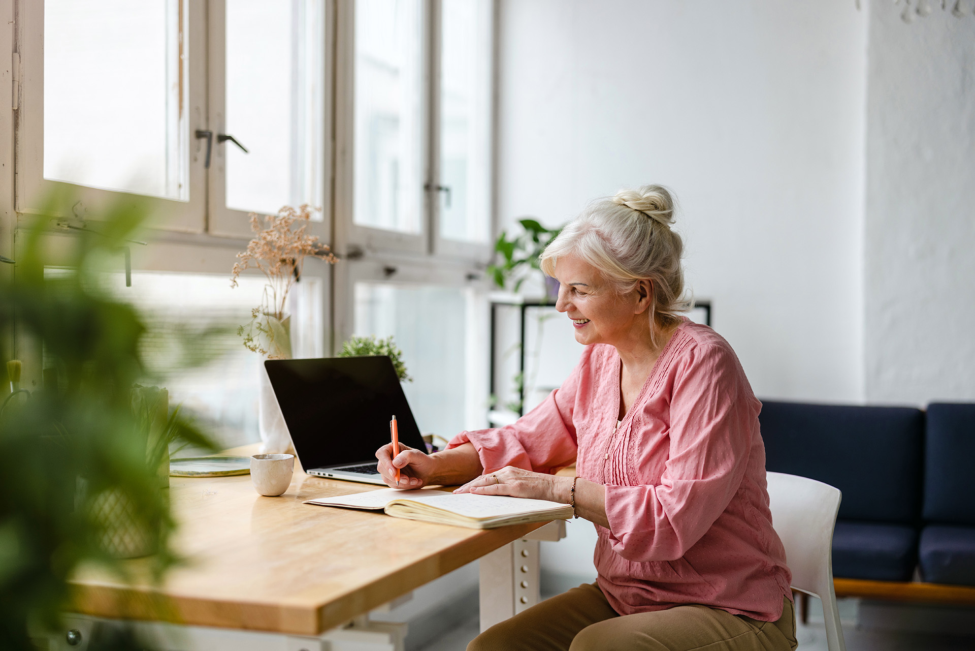 Smiling older woman with glasses sitting on a gray sofa, talking on a phone in a modern living room.