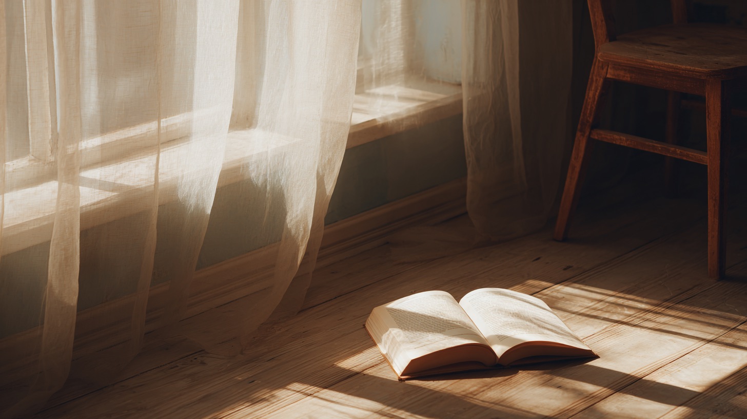 Livre ouvert posé sur un sol en bois éclairé par la lumière du soleil à travers un rideau transparent, avec une chaise en bois en arrière-plan.