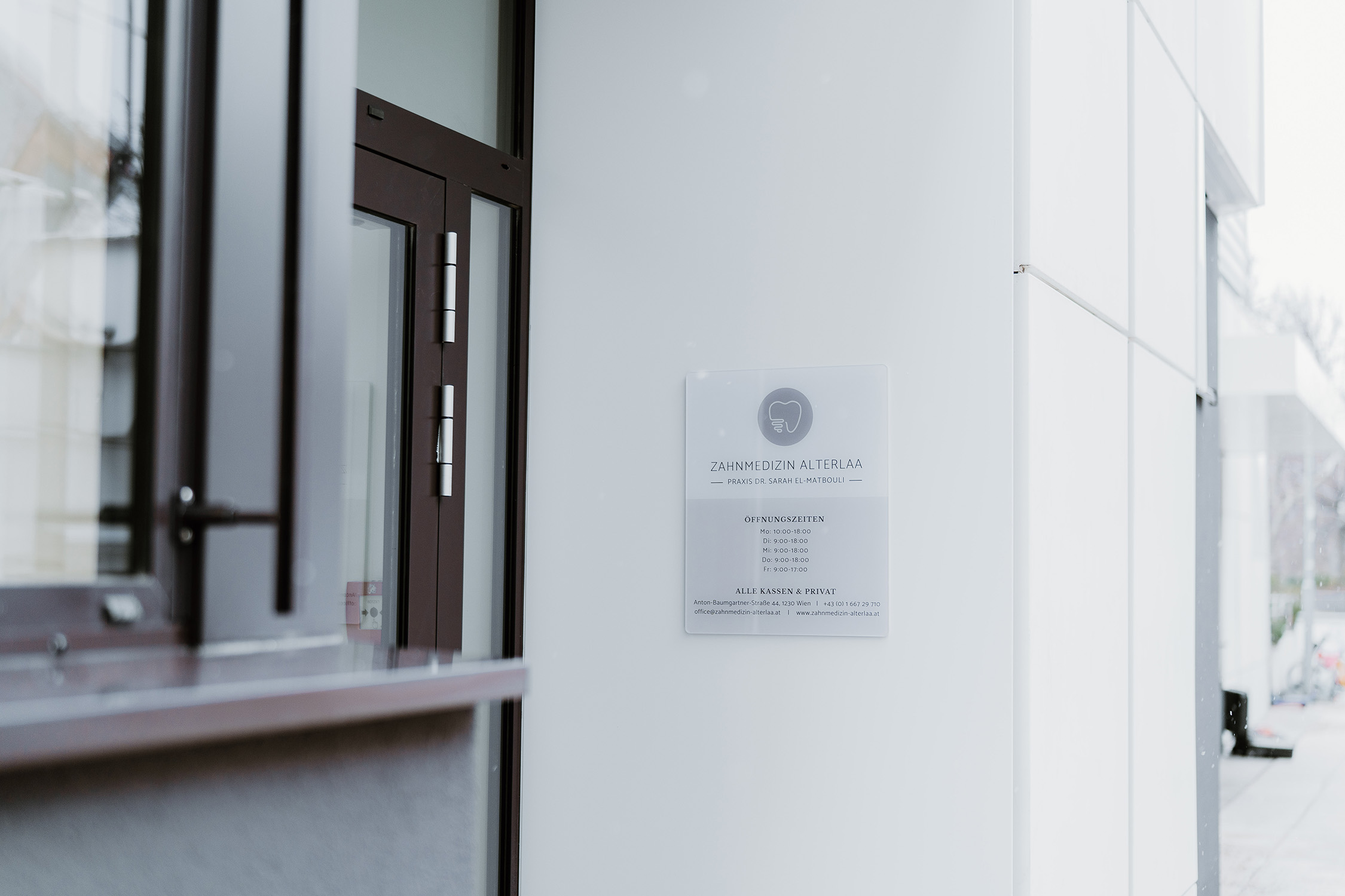Schild der Zahnarztpraxis Zahnmedizin Alterlaa neben brauner Glastür an weißer Hauswand.