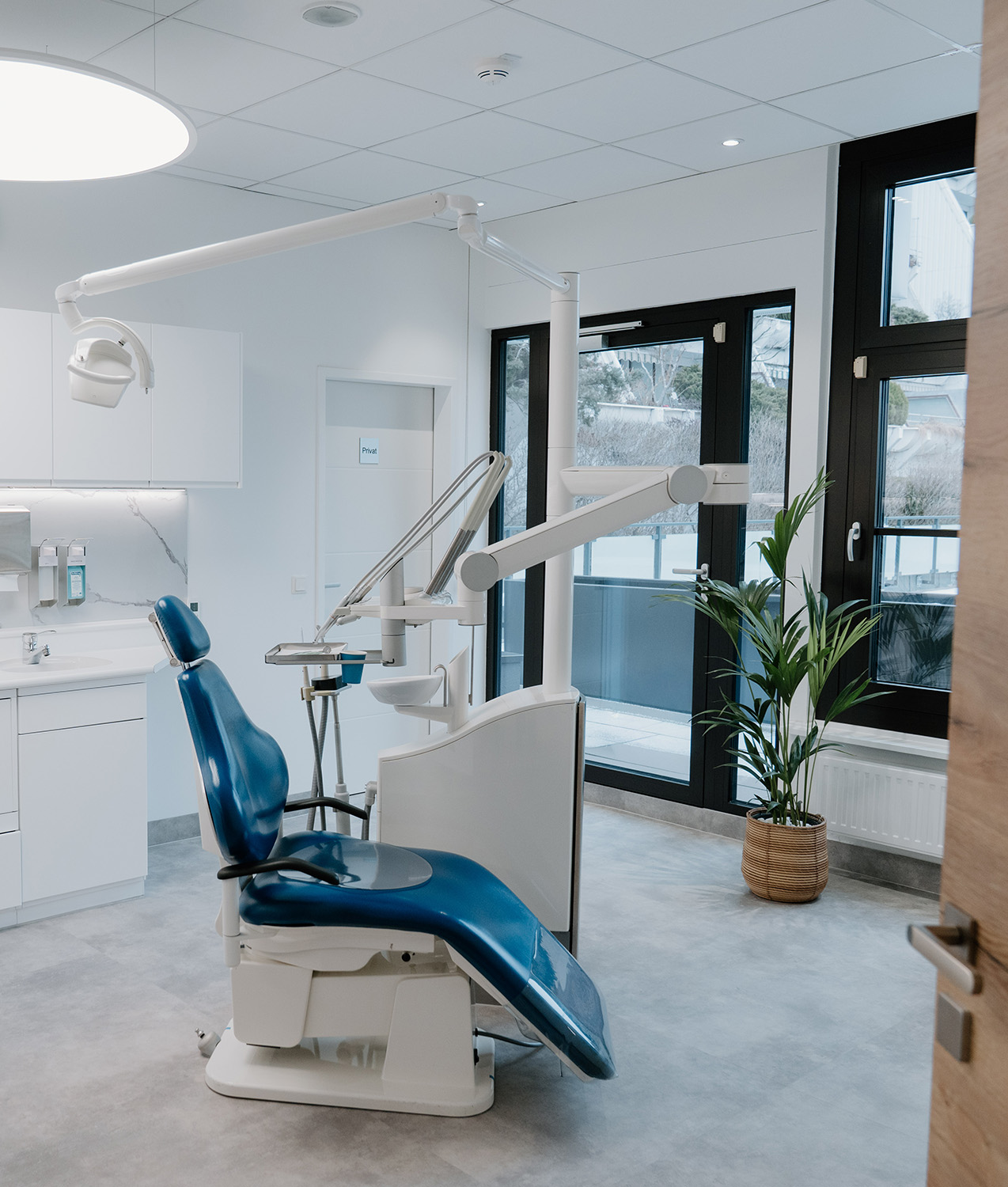 Moderner Zahnarztstuhl mit blauer Polsterung in hellen Behandlungszimmer mit großen Fenstern und grüner Pflanze.
