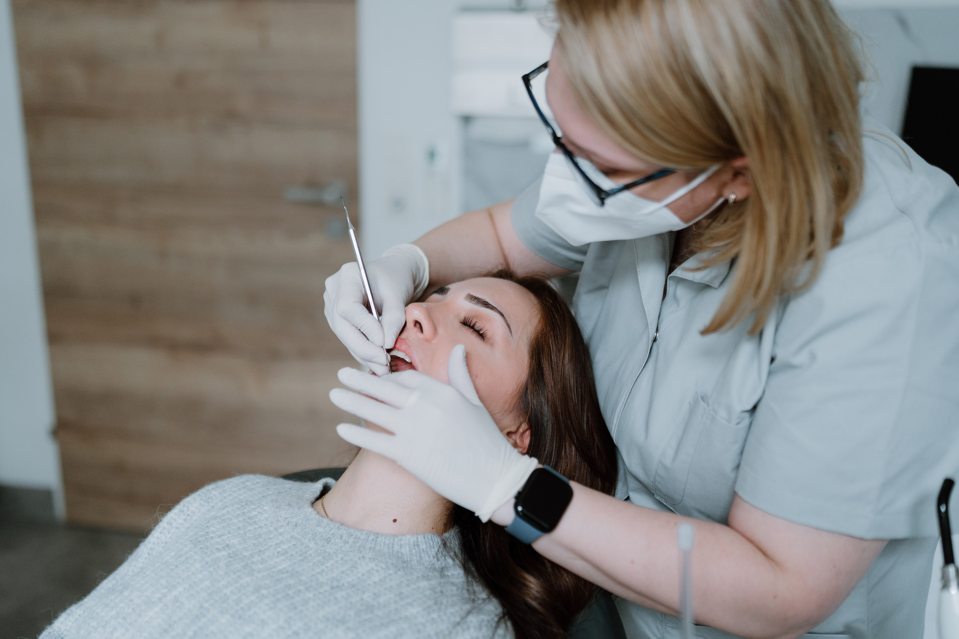 Eine Zahnärztin mit Maske und Handschuhen untersucht die Zähne einer entspannten Patientin in einer Zahnarztpraxis.