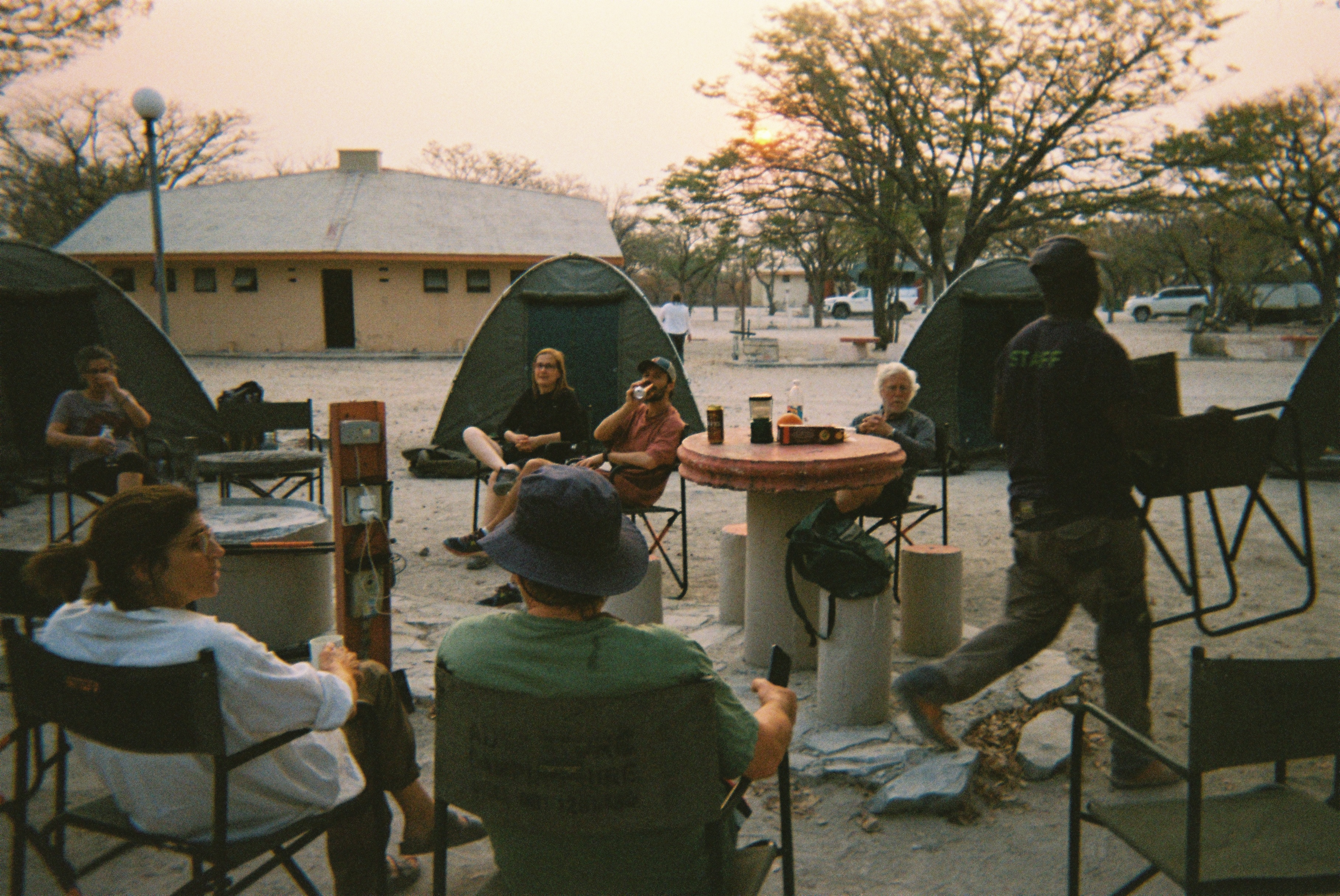 Group of people sitting in camping chairs around a round table with tents and trees in the background during sunset.