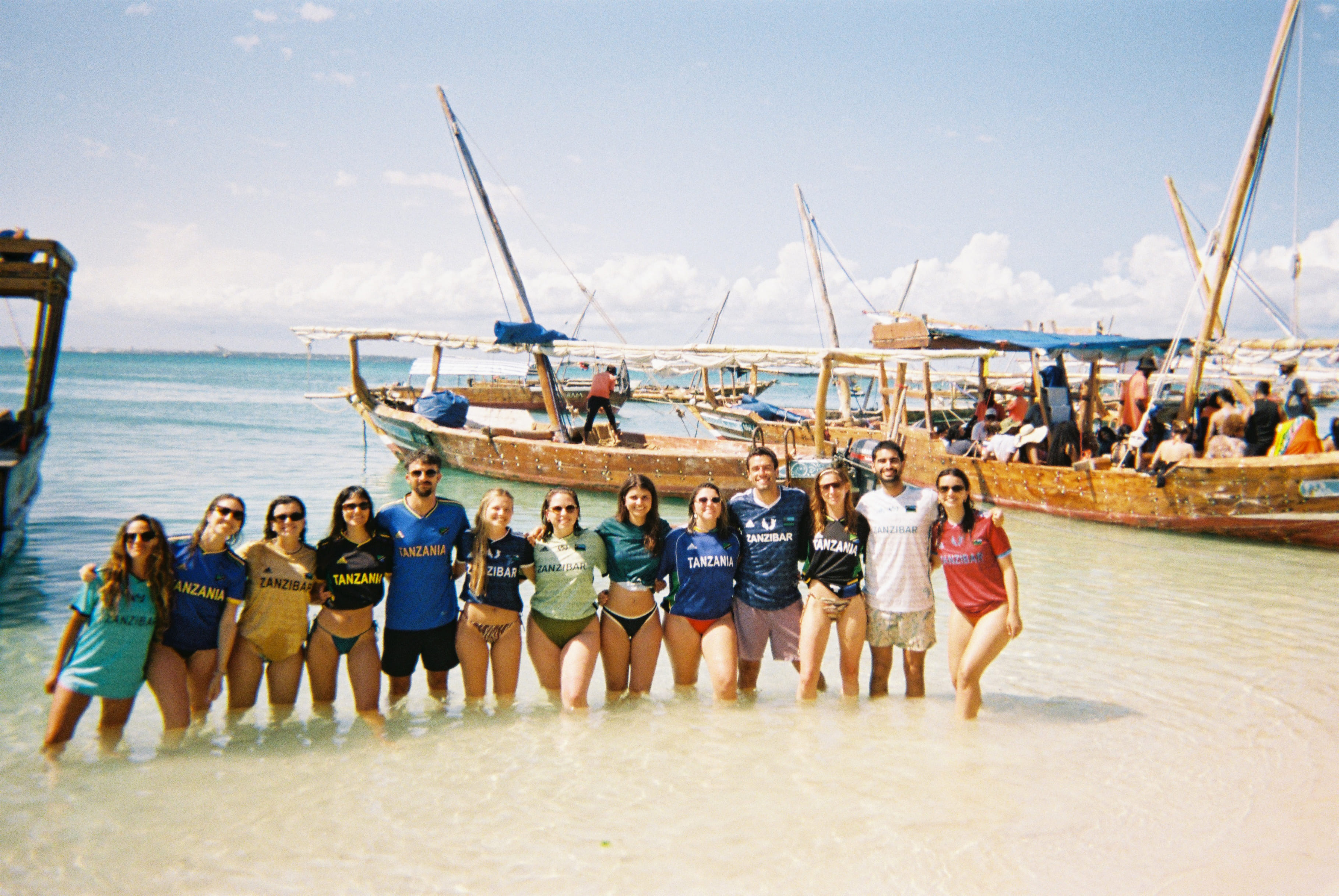 Group of people standing in shallow ocean water with traditional wooden boats in the background under a partly cloudy sky.