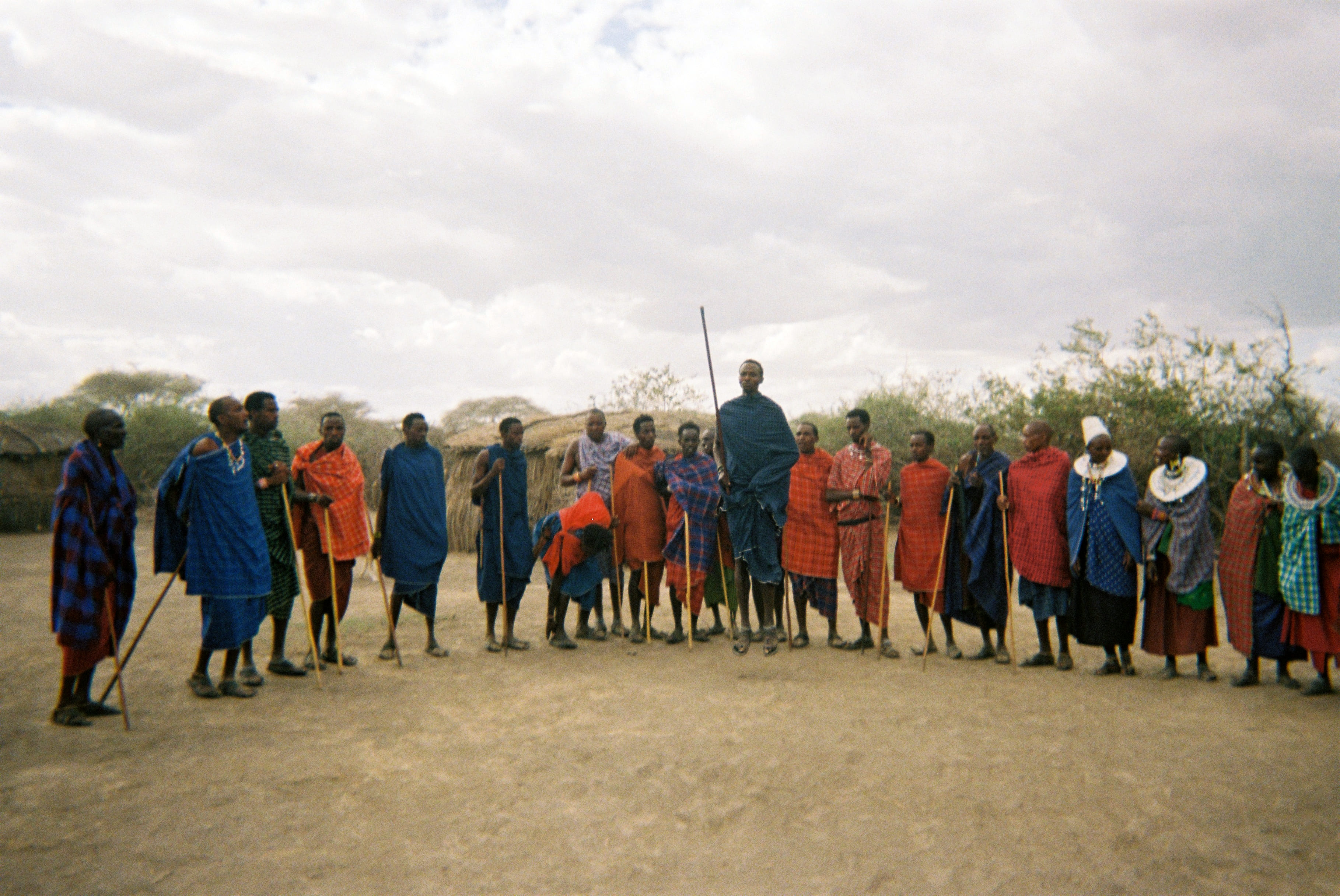 Group of Maasai people standing outdoors wearing traditional colorful shukas and holding walking sticks under a cloudy sky.