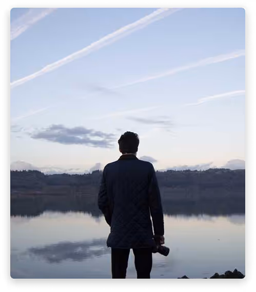 Man holding a camera stands facing a calm lake with hills in the background under a sky with scattered clouds and contrails.
