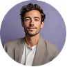 Headshot of a confident man with short curly hair and a light beard wearing a beige blazer and white shirt against a purple background.