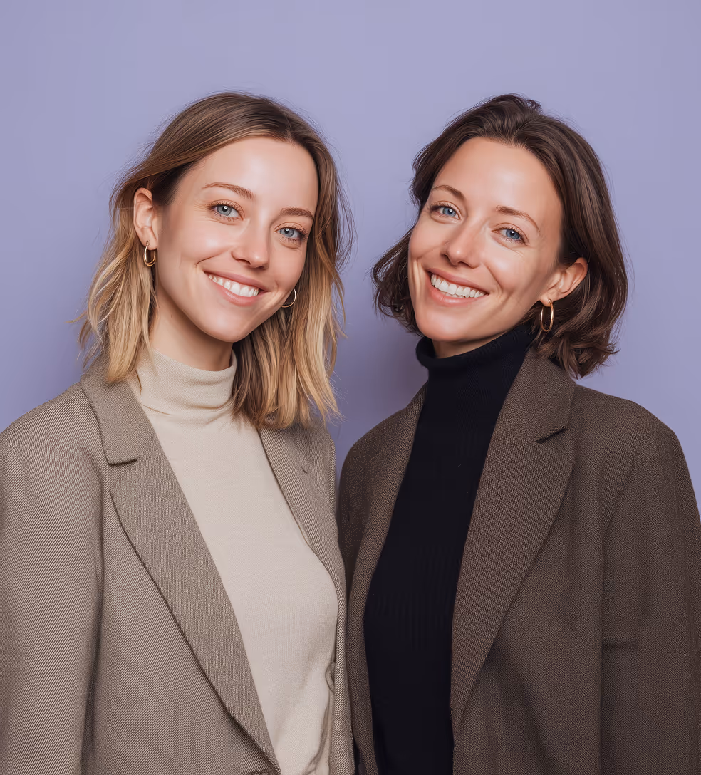 Two smiling women wearing blazers, one in beige and the other in dark brown, standing against a lavender background.