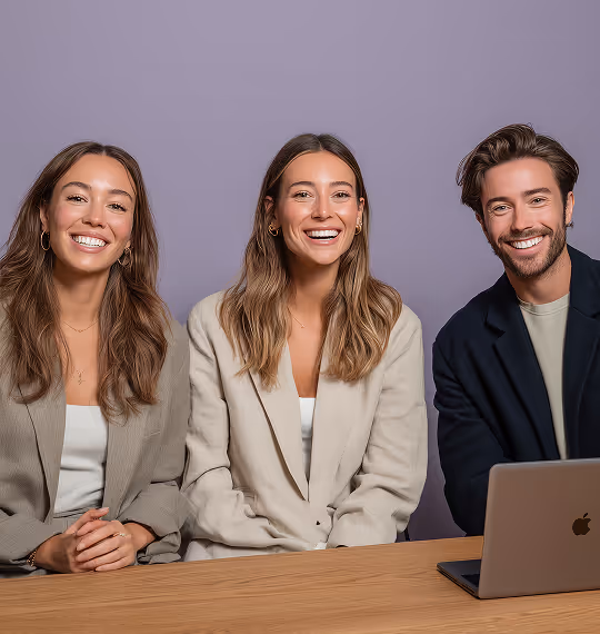 Three smiling young professionals sitting side by side at a table with a laptop in front of them against a purple background.
