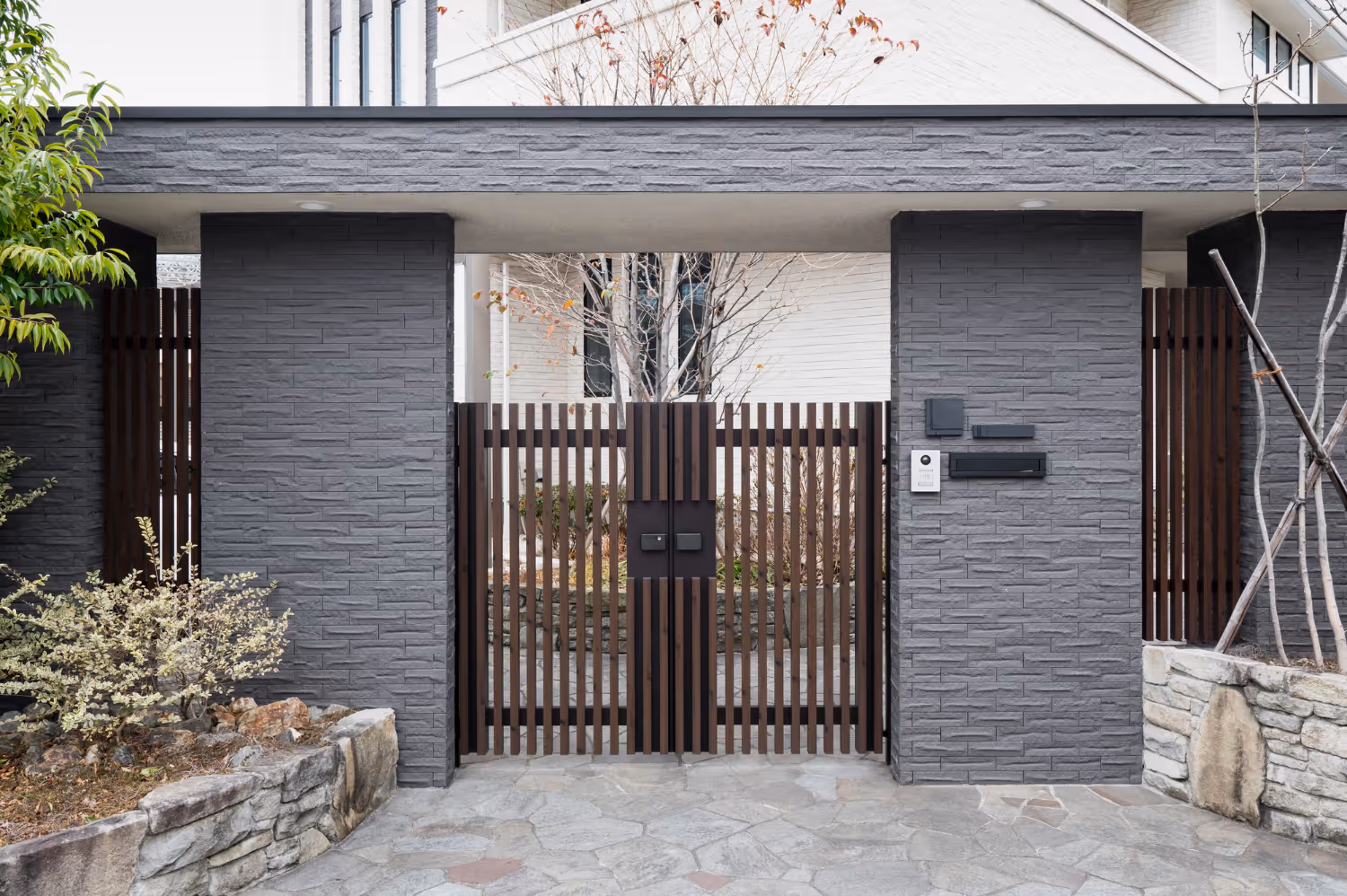 Modern house entrance with dark gray brick walls, a wooden slatted gate, and stone pavement.
