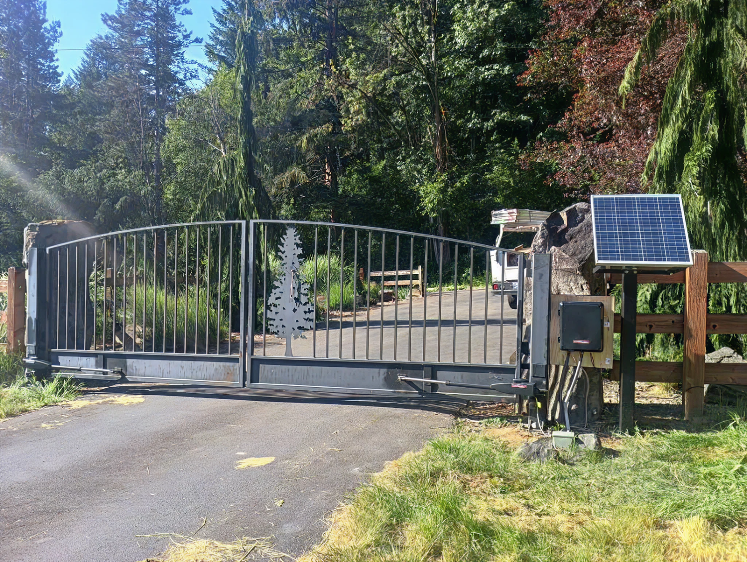 Closed metal gate with a tree silhouette design, a solar panel, and a mailbox next to a paved road surrounded by trees.