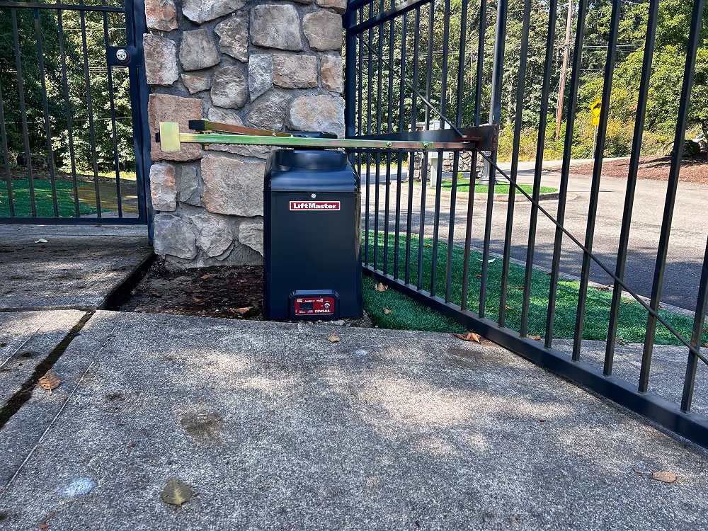 Close-up of a LifMaster automatic gate opener attached to a stone pillar and black metal gate on a concrete driveway.