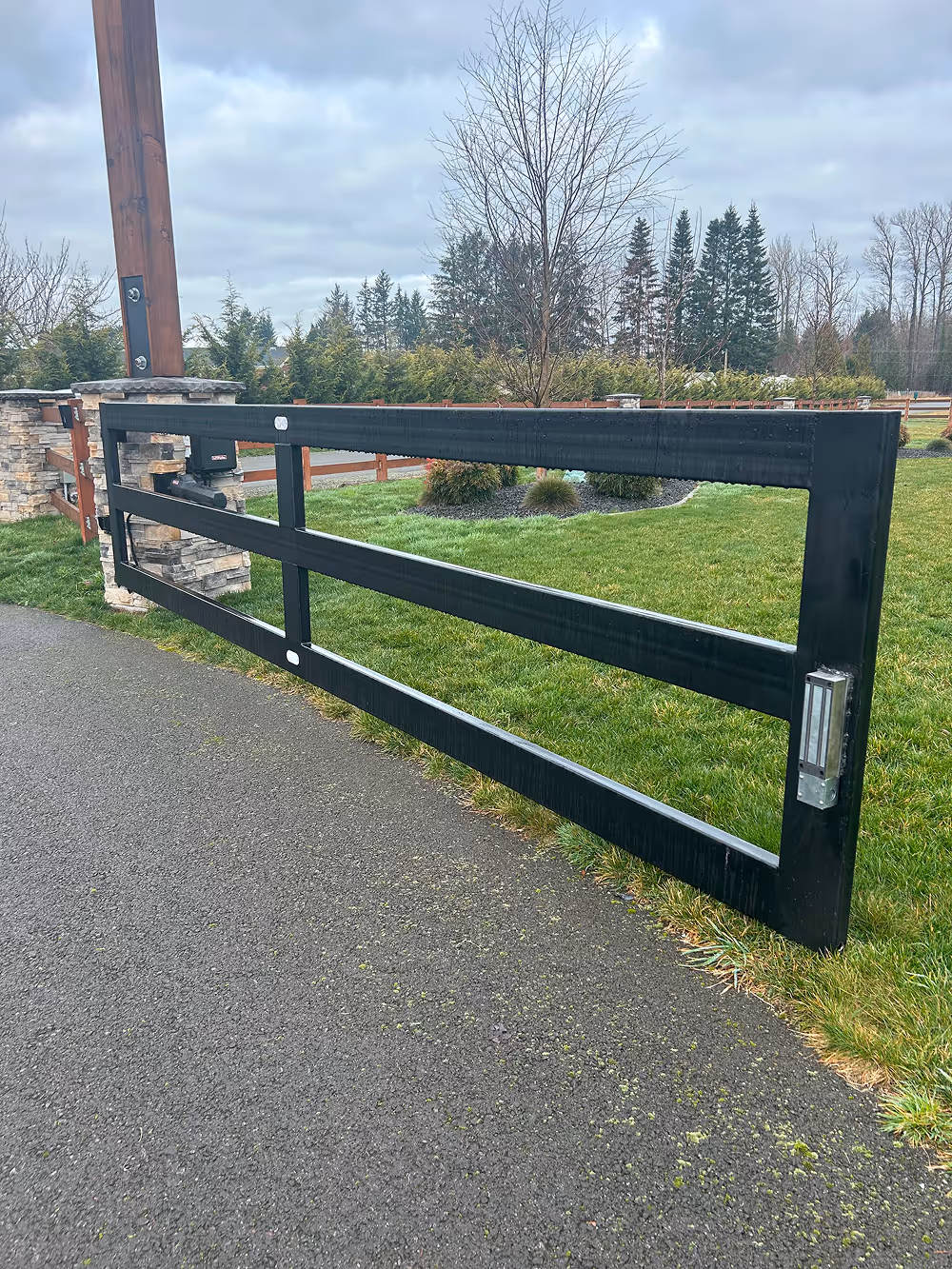 Black metal driveway gate swung open on an asphalt driveway with green lawn and trees in the background.