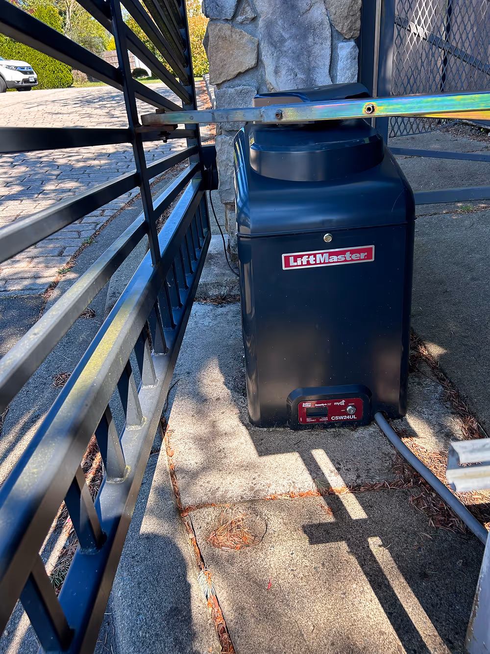 Close-up of a LiftMaster automatic gate opener attached to a black metal gate and stone pillar on a concrete driveway.