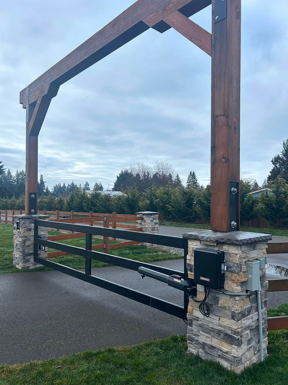 Automated black metal and wood driveway gate with stone pillars and wooden overhead frame in a rural setting.