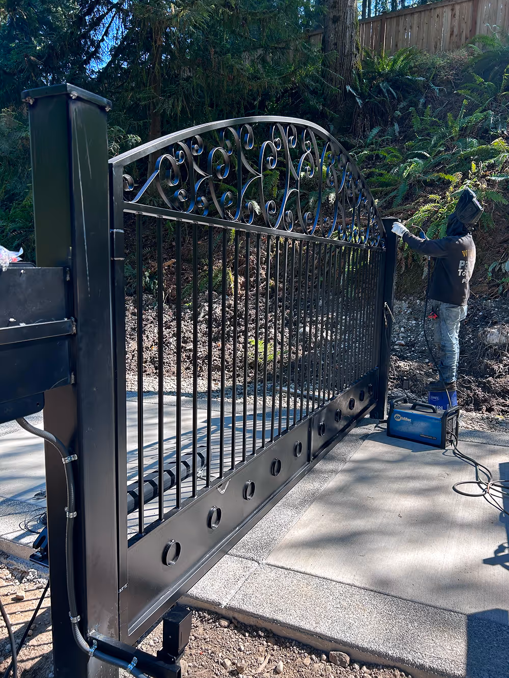 Person welding a decorative black metal gate outdoors near a wooded area.