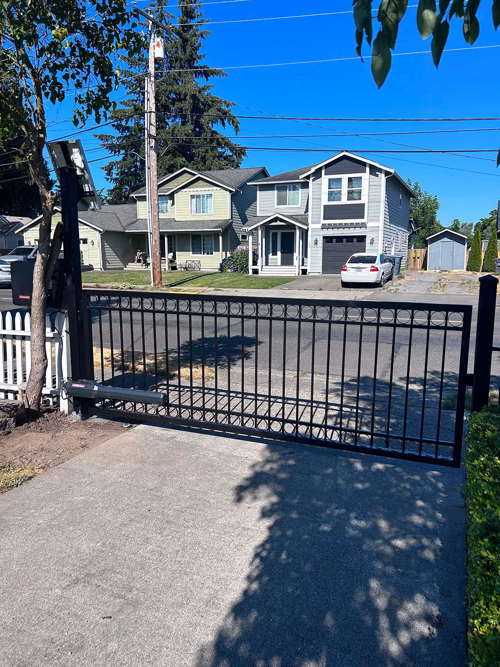 Black metal driveway gate with circular decorative elements, partially open, in front of a suburban street with houses and parked cars under a clear blue sky.