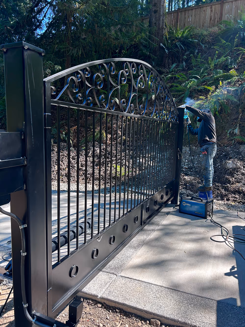 Person welding a large black metal gate outdoors next to a wooded area.