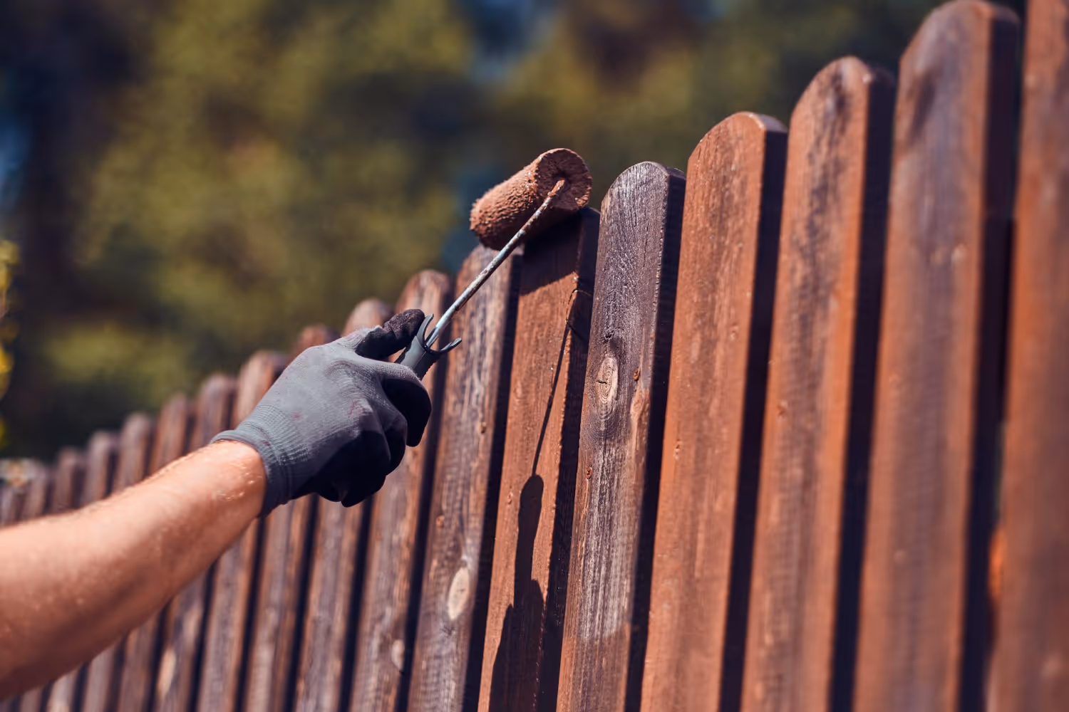 Person wearing black glove painting a wooden fence with a paint roller in an outdoor setting.