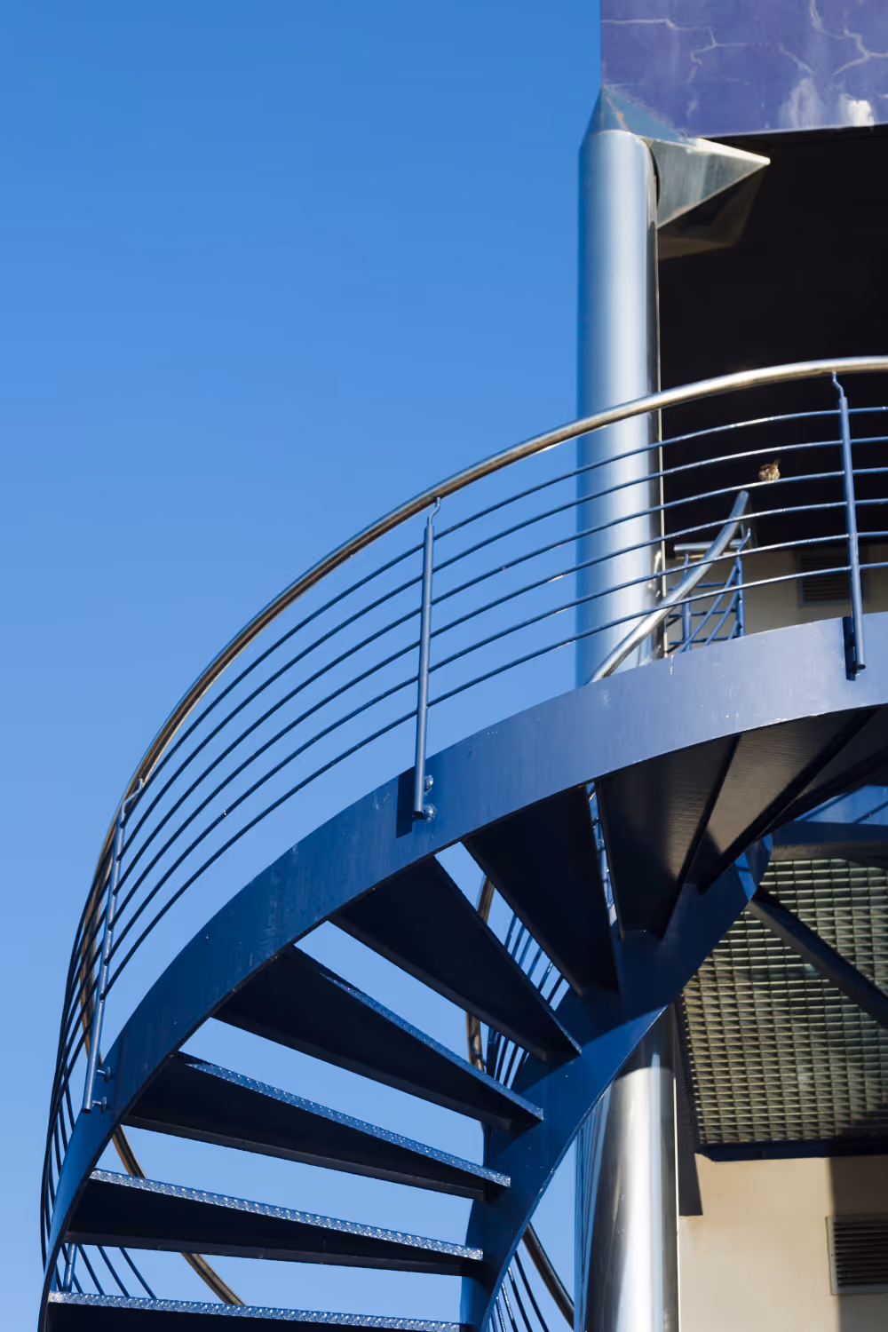 Close-up of a modern blue spiral staircase with metal railings against a clear blue sky.