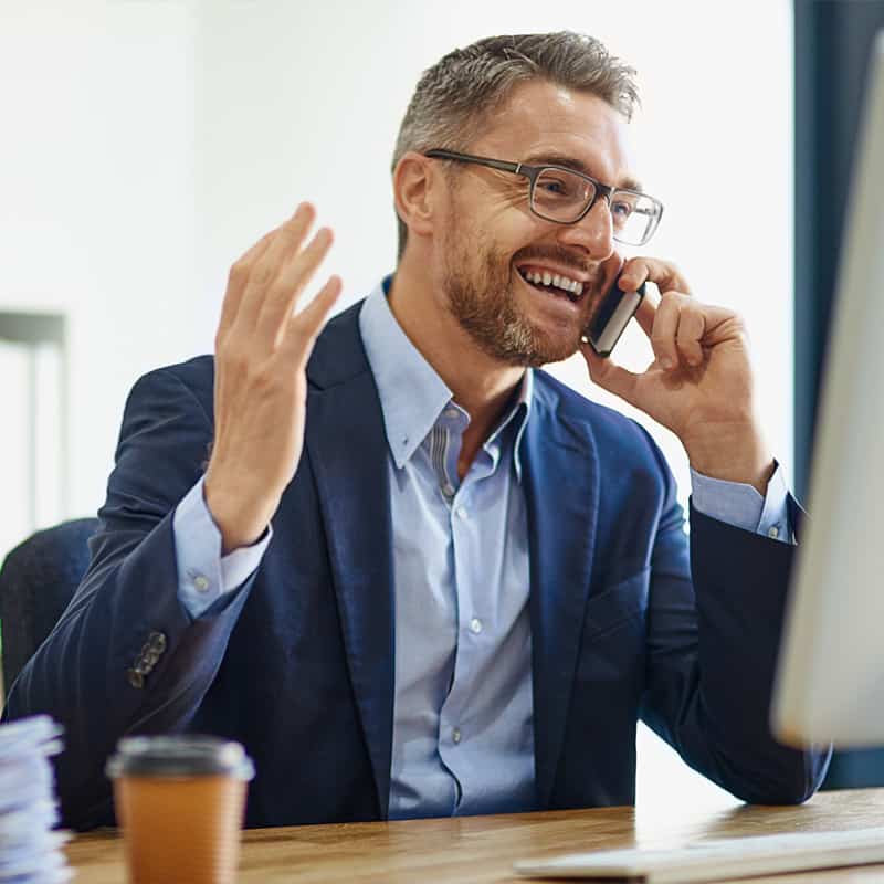 Smiling businessman in glasses talking on a smartphone at his desk in an office.