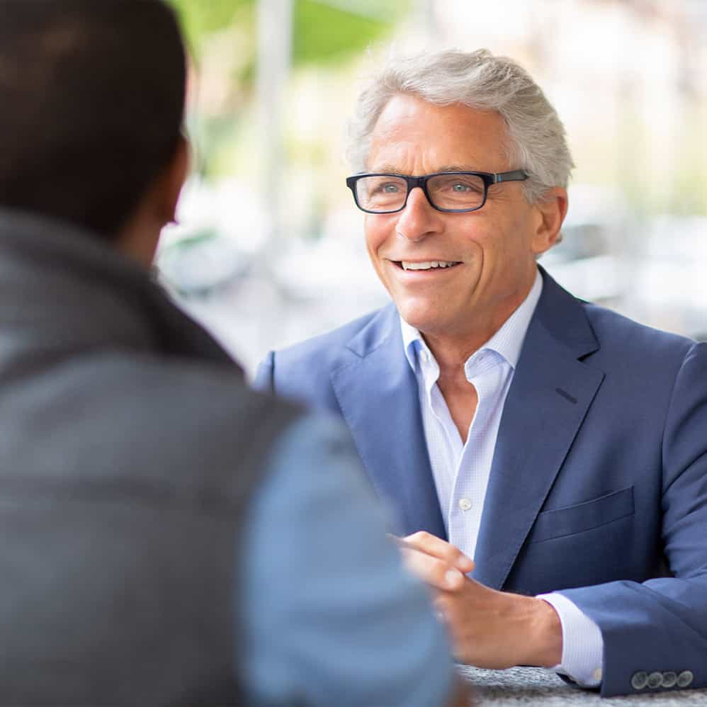 Smiling older man with gray hair and glasses engaged in conversation with a younger person.
