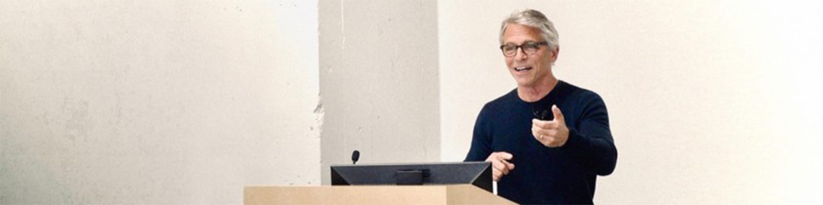 Man with gray hair and glasses speaking at a podium in a minimal setting.