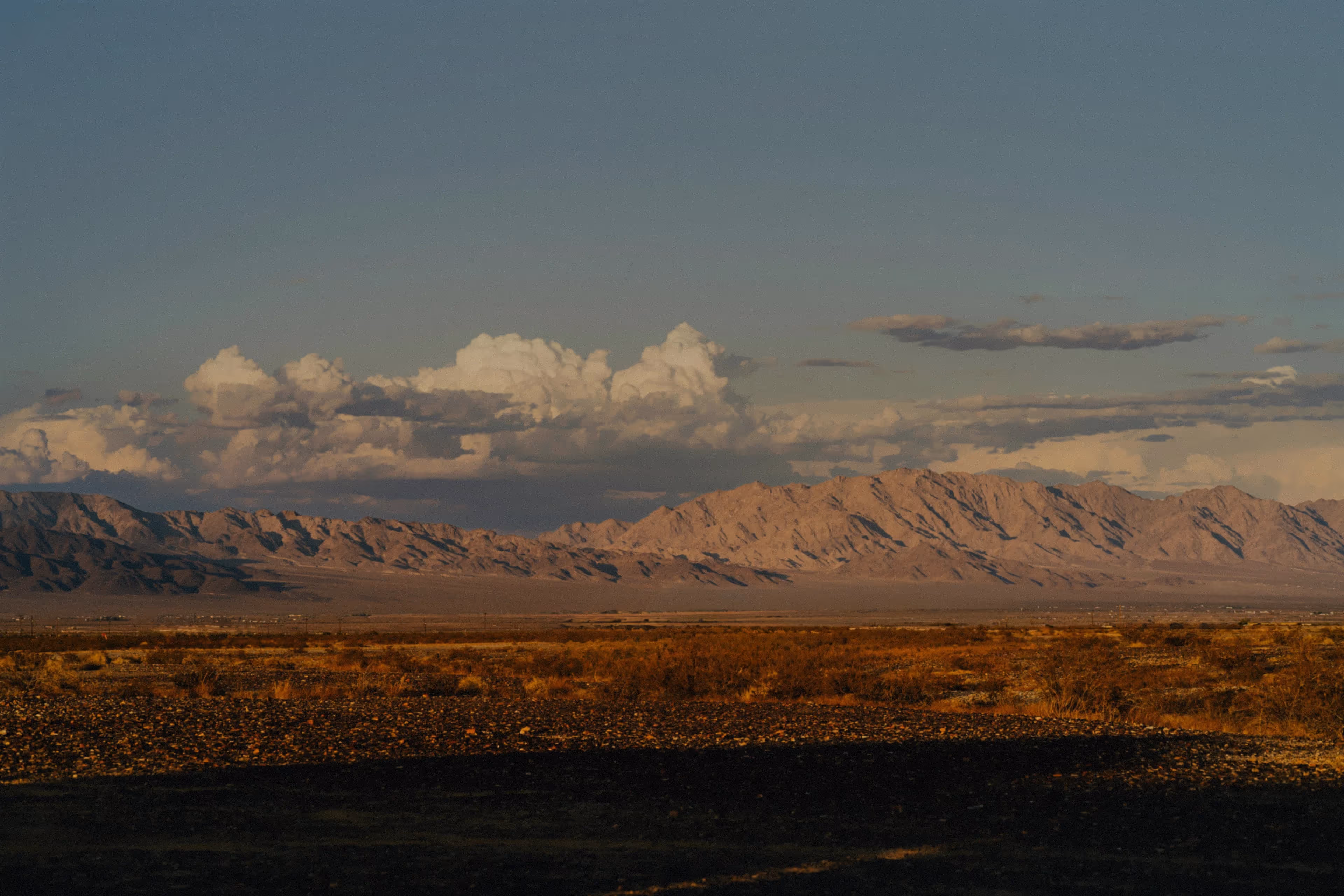 Desert landscape with rocky terrain and sparse vegetation under a sky with large clouds and distant mountains illuminated by warm light.