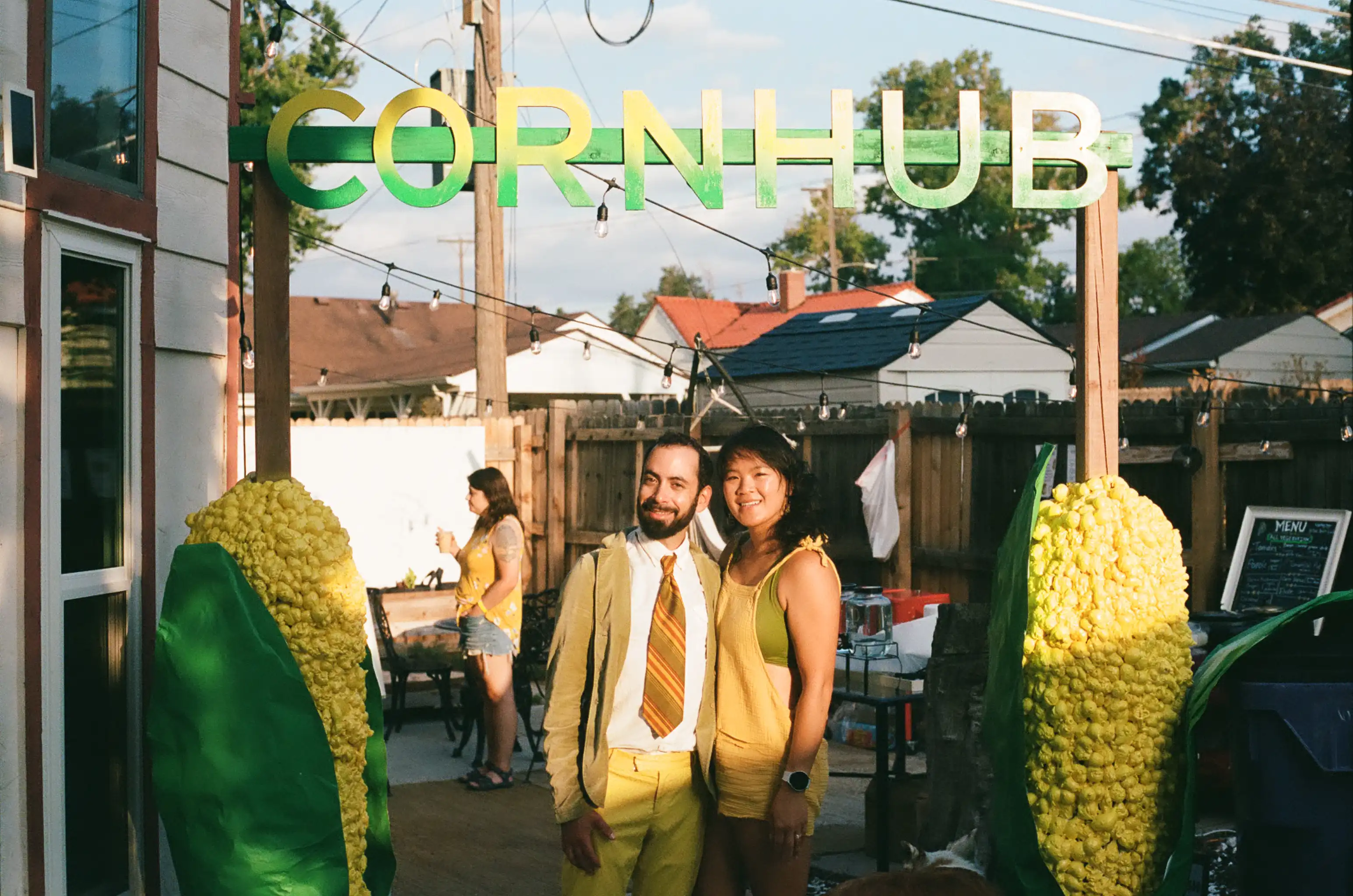 Two people smiling and posing under a green and yellow sign that says CORNHUB, with large corn sculptures on either side, at an outdoor event.