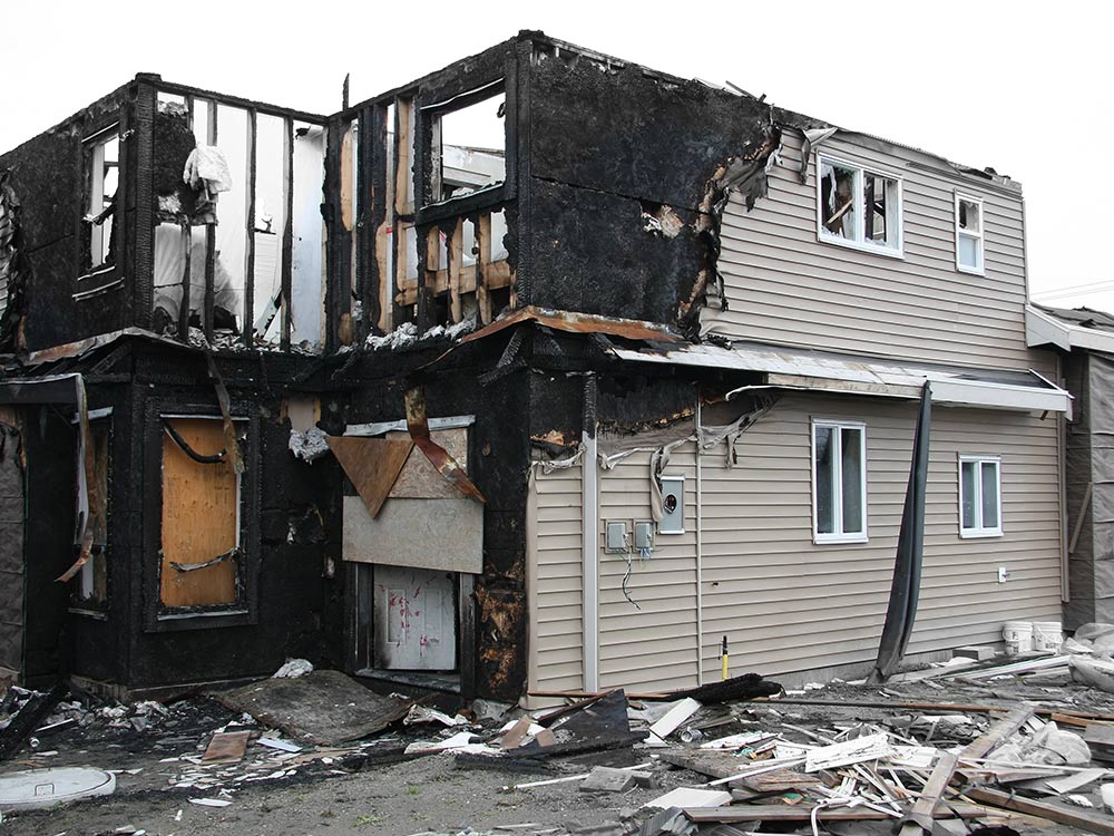 Two-story house severely damaged by fire, with charred black walls on the left side and intact beige siding on the right, broken windows, boarded-up door, and debris scattered around the building exterior.