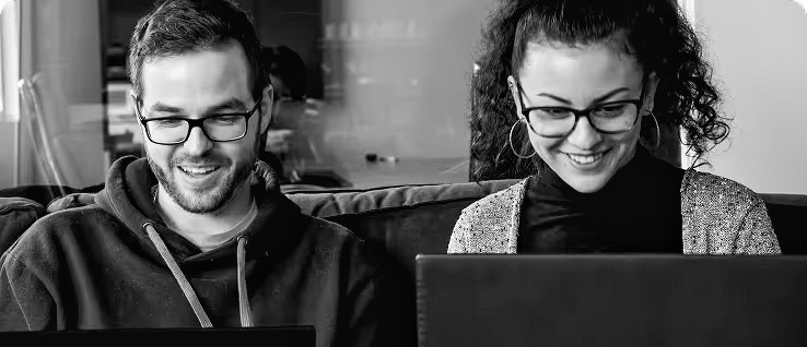 Deux personnes portant des lunettes, travaillant côte à côte sur des ordinateurs portables et souriant.