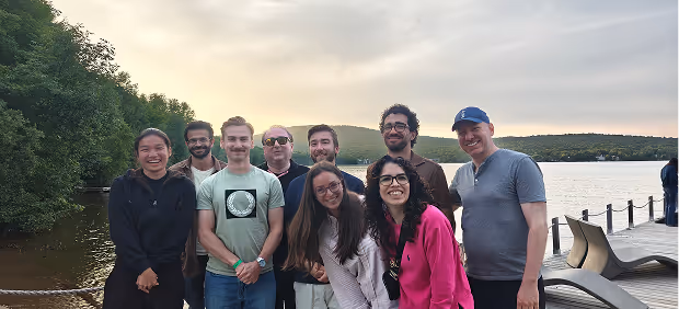 Groupe de huit personnes souriantes posant près d'un lac avec des arbres et un quai au coucher du soleil.