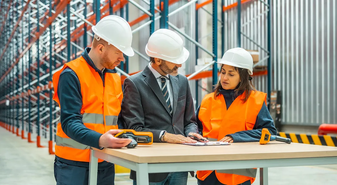 Three warehouse workers in hard hats and orange safety vests discussing documents on a table in an industrial storage facility.