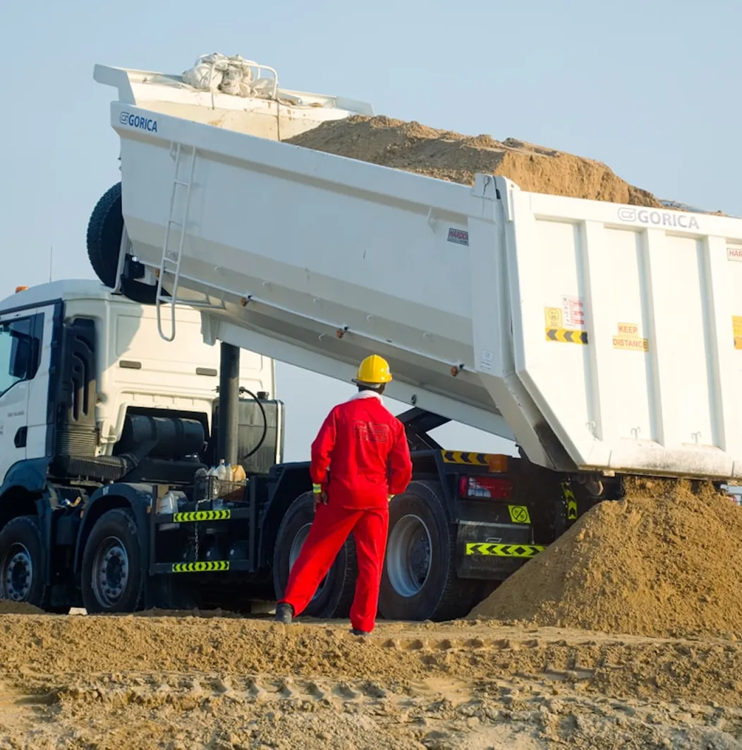 Construction worker in red coveralls and yellow helmet standing near a white dump truck unloading sand at a construction site.