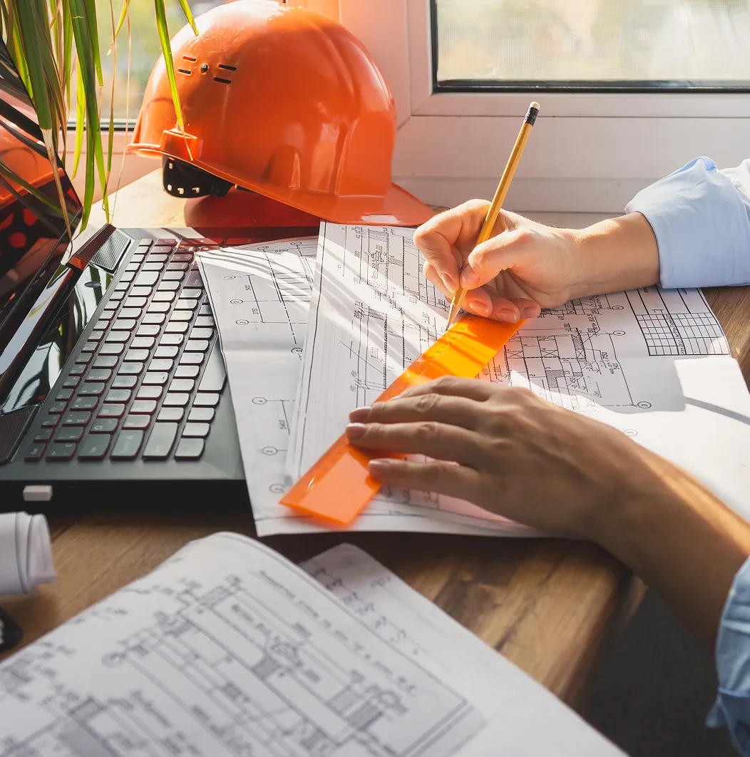 Person drawing precise lines on architectural blueprints with an orange ruler and pencil next to a laptop and orange safety helmet.