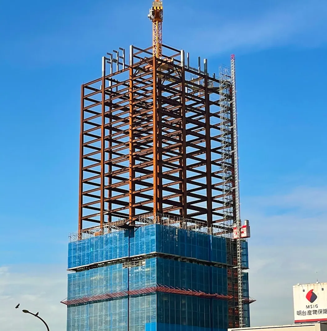 High-rise building under construction with exposed steel framework above a glass-covered lower structure against a clear blue sky.