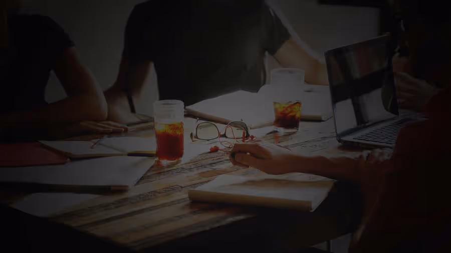 People sitting around a wooden table with notebooks, glasses of iced beverages, and a laptop during a meeting.