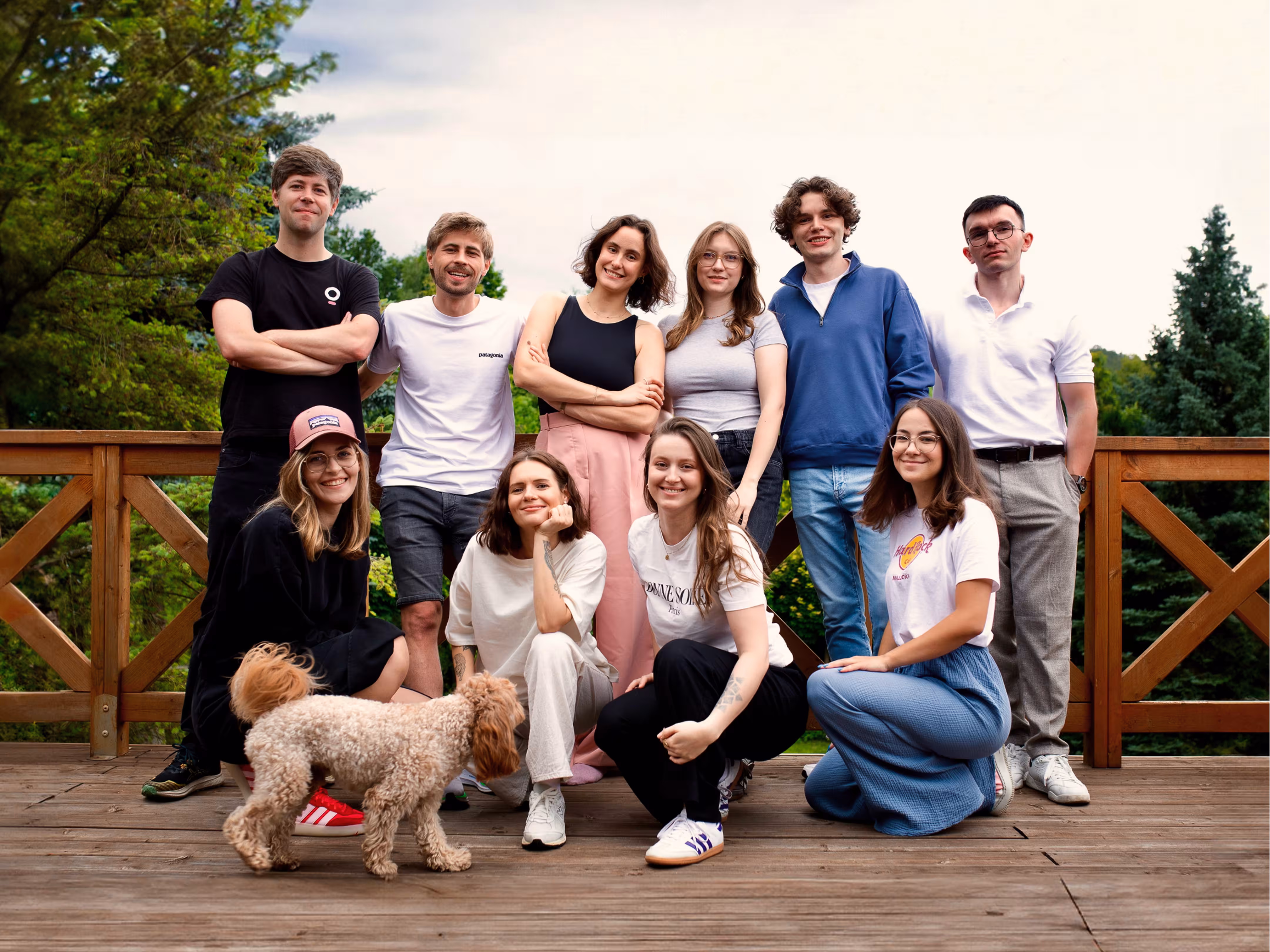 Group of ten young adults posing together on a wooden deck with trees in the background and a small curly-haired dog in front.