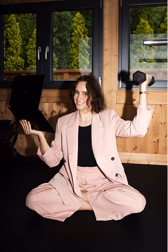 Woman in pink suit sitting cross-legged indoors, holding a laptop in one hand and a dumbbell in the other, smiling.