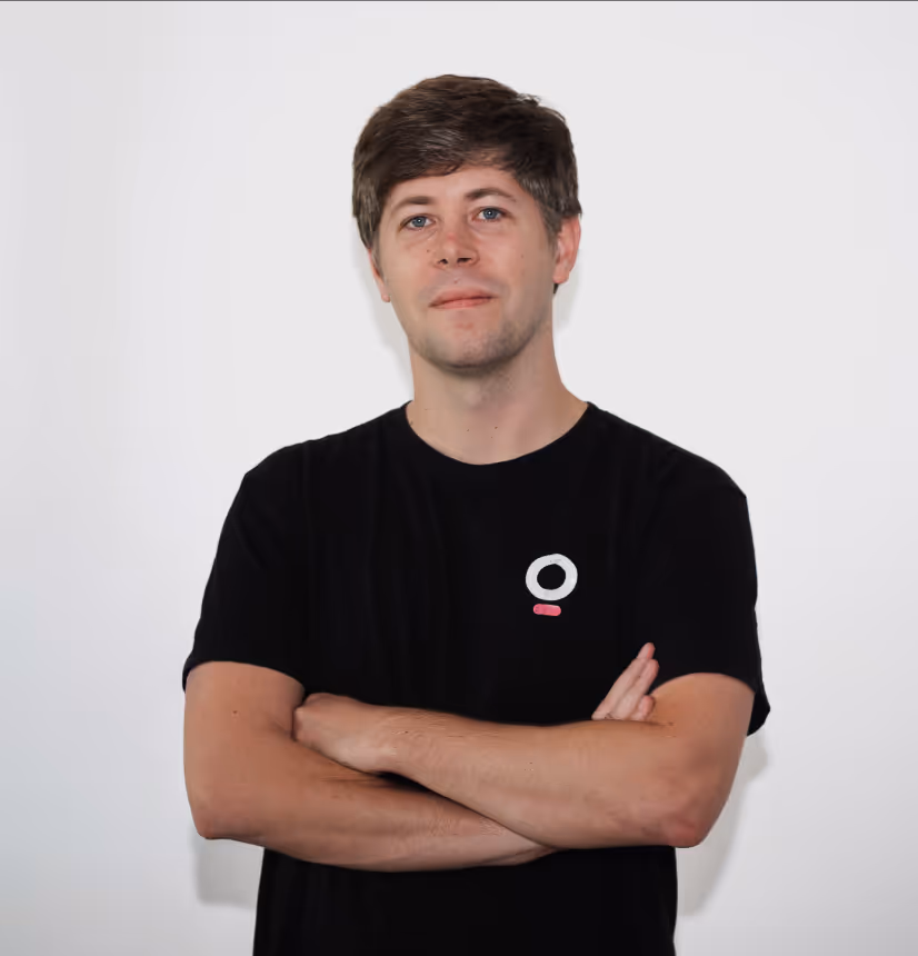 Young man with brown hair wearing a black t-shirt with a small white and pink logo, standing against a plain white background with arms crossed.