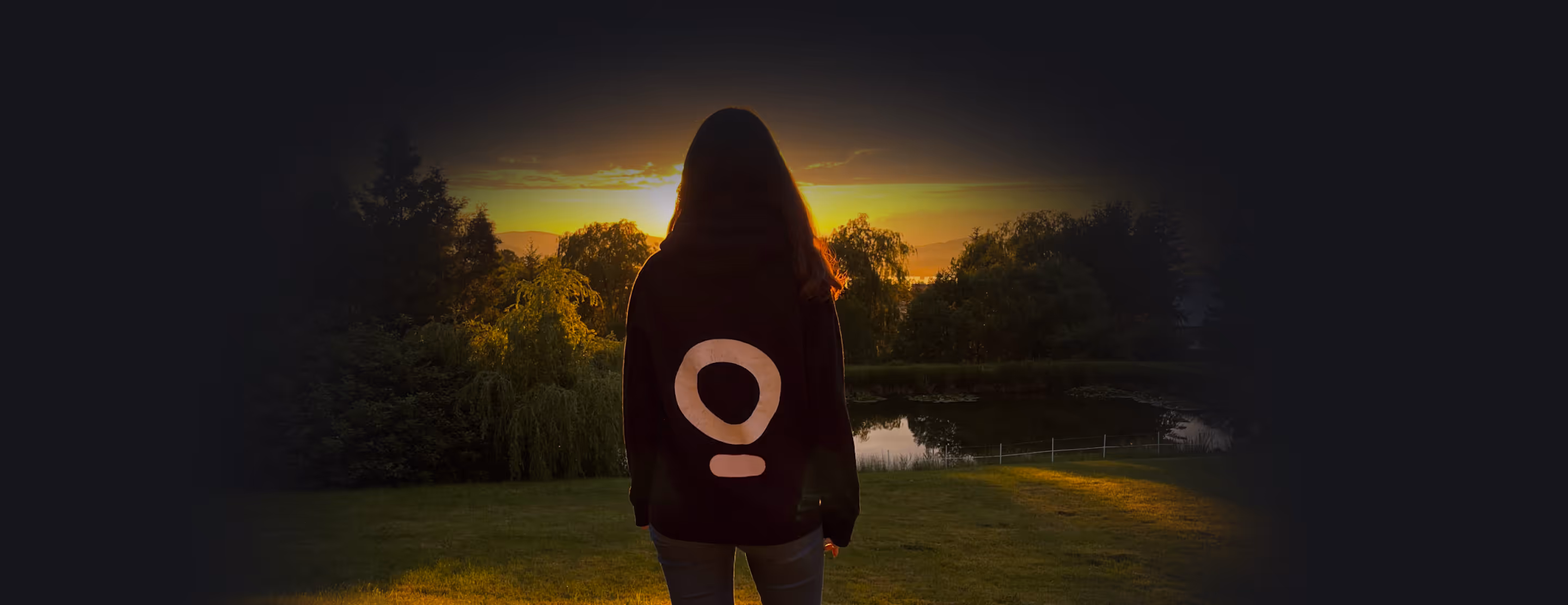 Person with long hair standing outdoors at sunset, facing a pond and surrounded by trees.