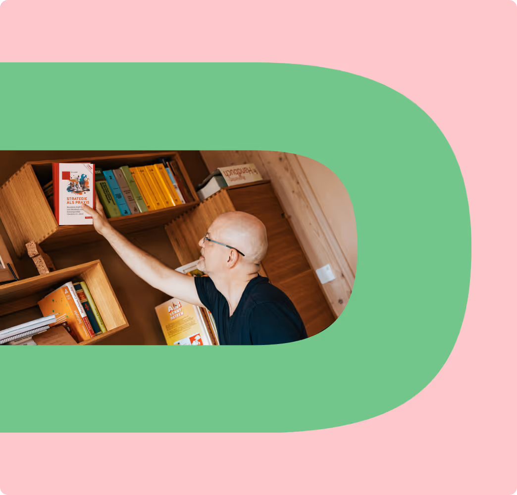 Bald man in glasses reaching for a book titled 'Strategie als Praxis' on a wooden shelf.