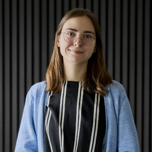 Junge Frau mit Brille, schwarzem Kleid mit weißen vertikalen Streifen und blauem Cardigan vor einem schwarzen, vertikal gestreiften Hintergrund.