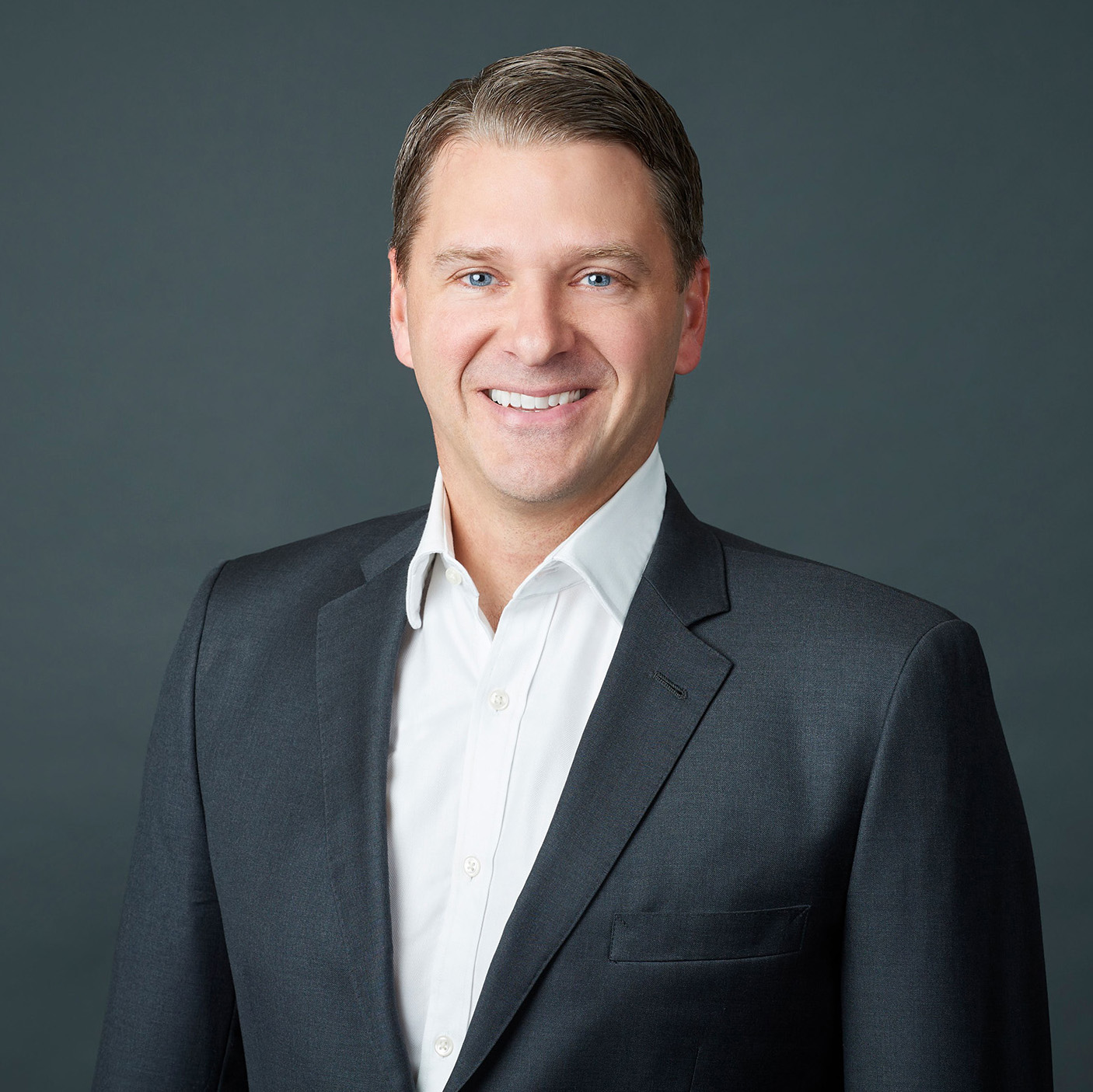 Smiling man with short brown hair wearing a dark gray suit jacket and white dress shirt against a gray background.