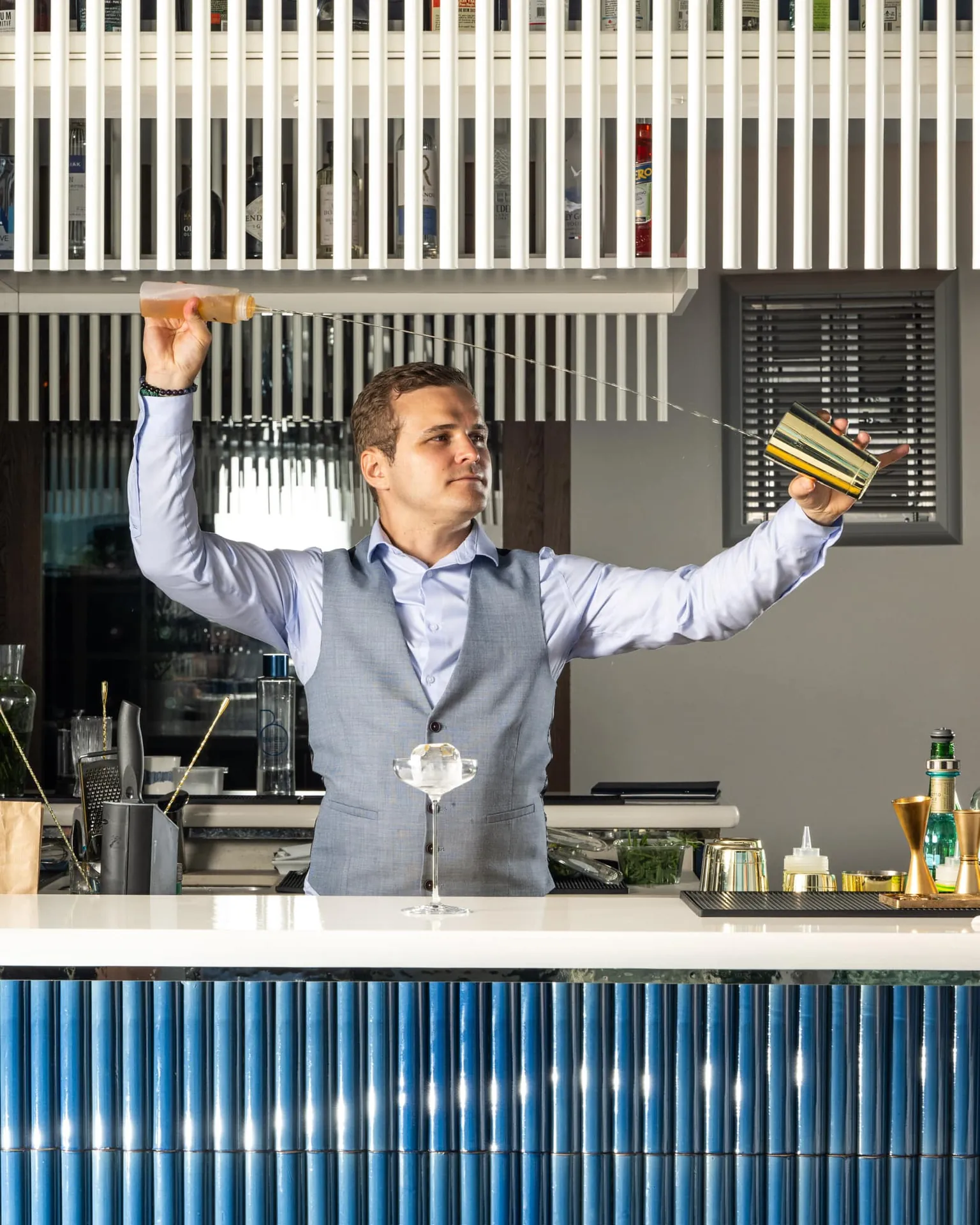 Victor Weber réalisant un throwing de cocktail derrière un bar en carrelage bleu