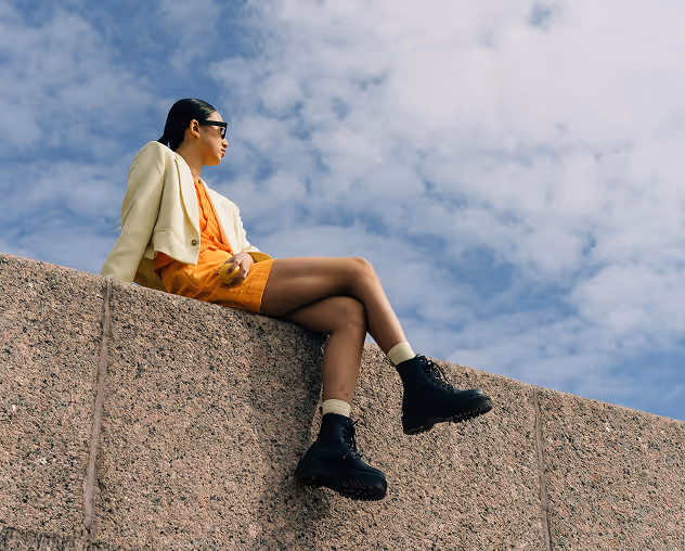 Man zit op een stoel met een bordeauxrood overhemd, groene broek en sieraden, pratend met een persoon naast hem.