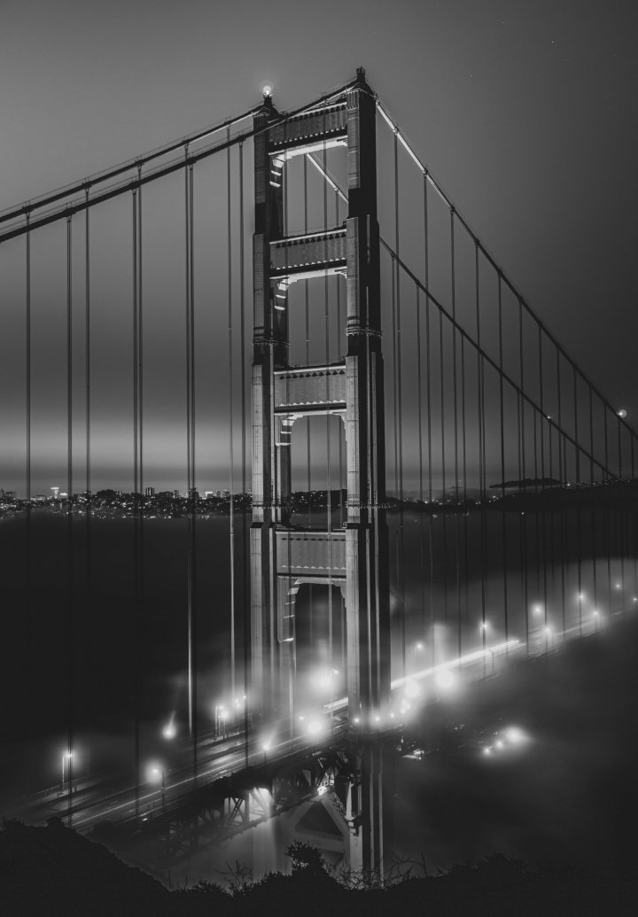 Black and white night view of the illuminated Golden Gate Bridge tower rising above fog with city lights in the background.